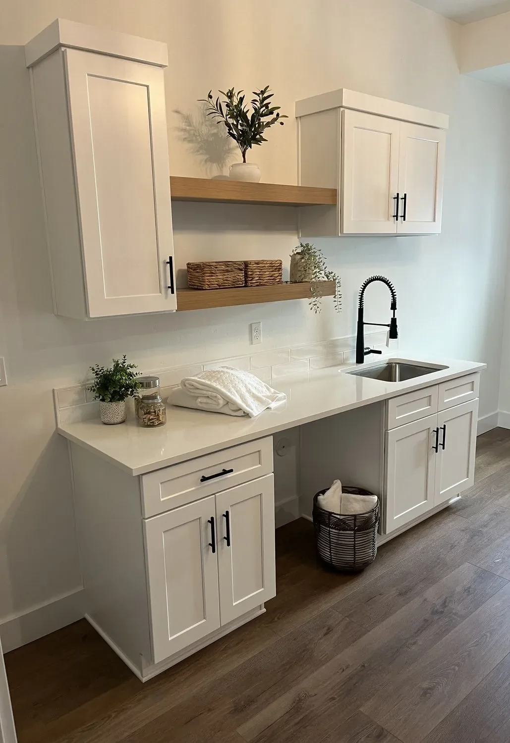 White shaker cabinets with stainless steel sink, floating wood shelves, and black hardware in laundry room